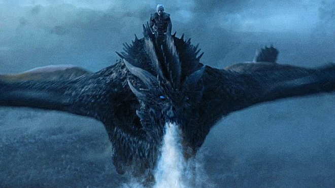 A dark, dramatic scene from Game of Thrones showing the Night King riding on the back of the ice dragon Viserion. The dragon's massive wings are spread wide against a stormy blue-gray sky as it breathes blue fire.