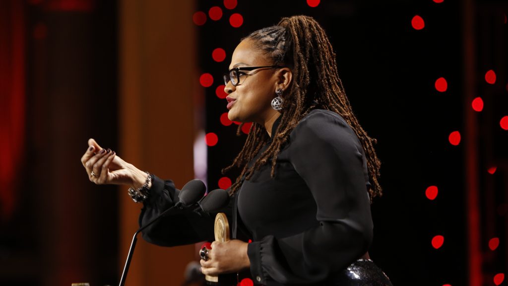 Ava DuVernay accepting an award on stage, speaking into a microphone while holding a trophy.