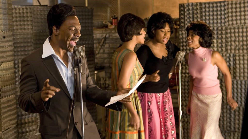 James 'Thunder' Early (Eddie Murphy) performing with backup singers Deena Jones (Beyoncé), Lorrell Robinson (Anika Noni Rose), and Effie White (Jennifer Hudson) in a scene from "Dreamgirls," singing into vintage microphones in a recording studio setting.