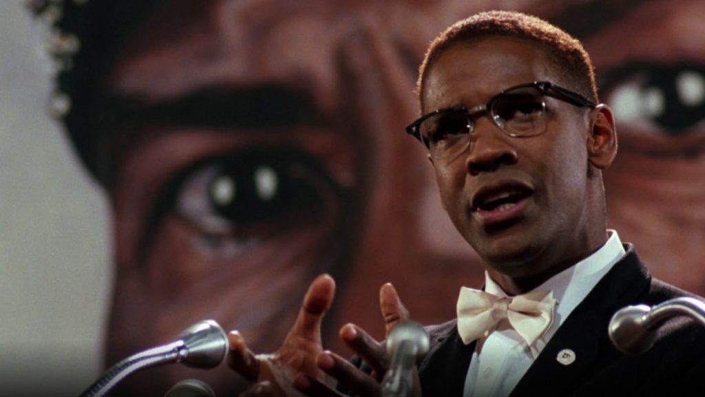 Denzel Washington as Malcolm X speaking at a podium wearing glasses and a bow tie, with a large portrait of Malcolm X visible behind him.