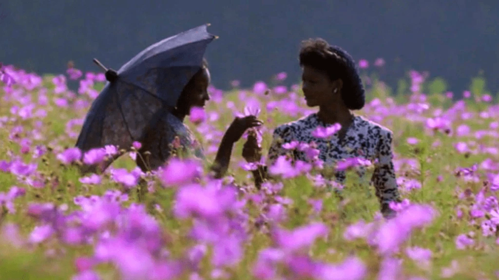 A scene from "The Color Purple" showing Celie (Whoopi Goldberg) and Shug (Margaret Avery) in a field of purple flowers, Celie holding an umbrella.