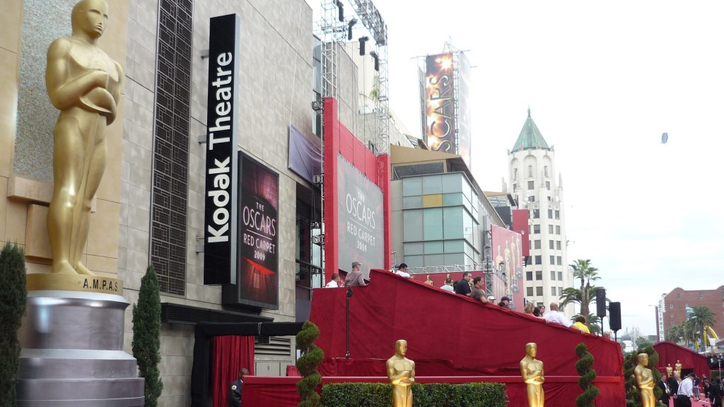 The exterior of the Kodak Theatre (now Dolby Theatre) decorated for the Academy Awards, featuring Oscar statues and red carpet preparations along Hollywood Boulevard.