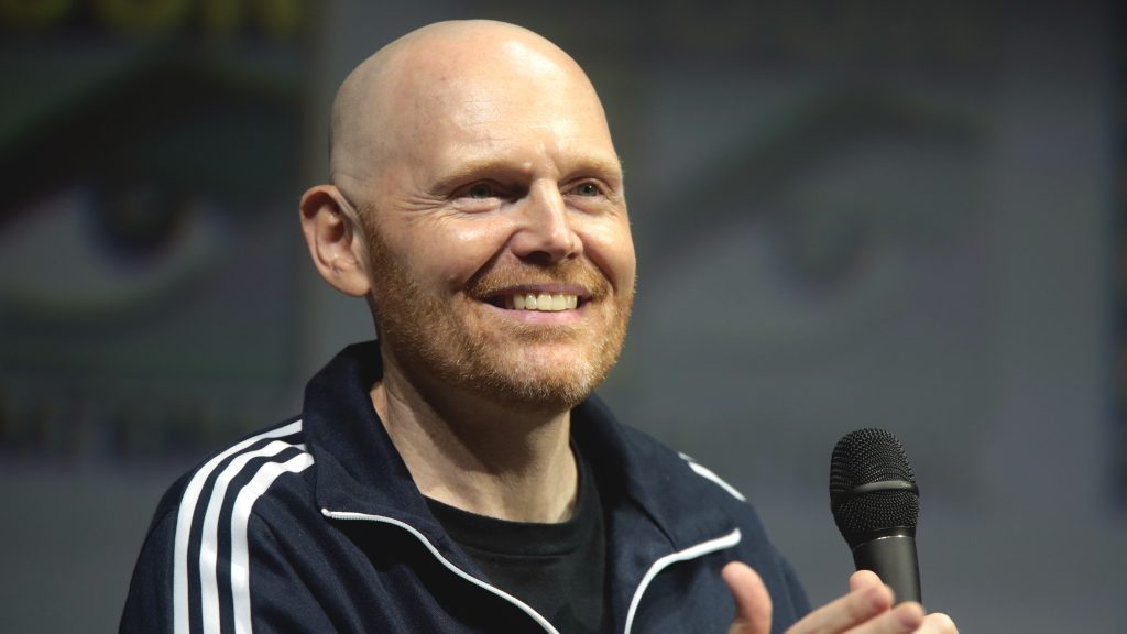 Comedian Bill Burr smiling while holding a microphone, wearing a dark jacket with white stripes.