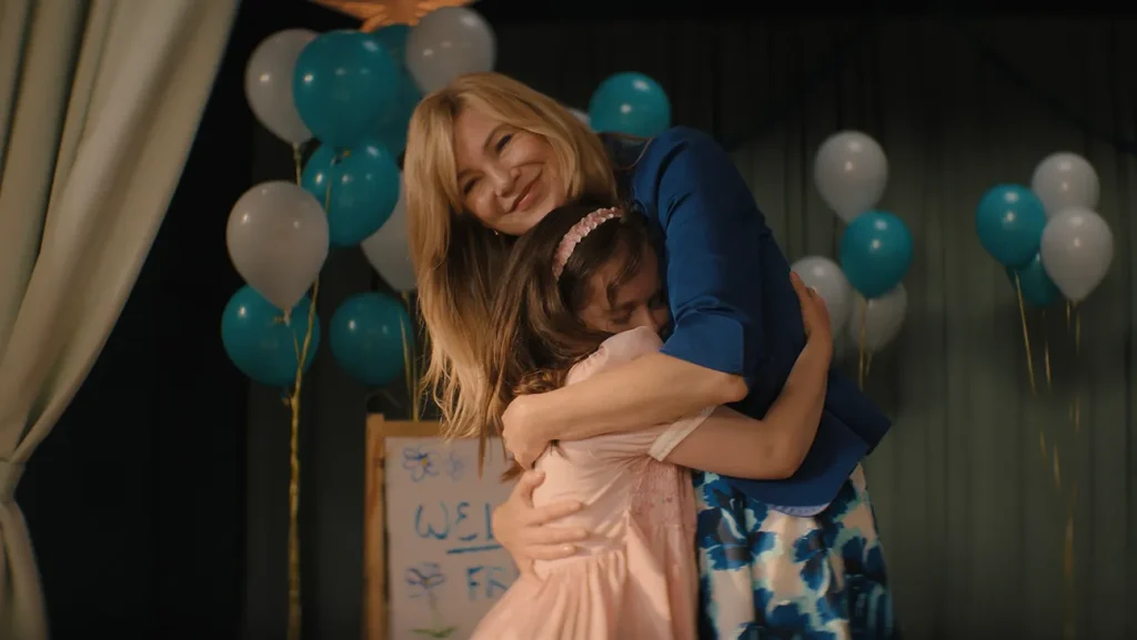 Ellen Pompeo in a blue outfit hugging Imogen Faith Reid in a pink dress, with blue and white balloons in the background, from "Good American Family."