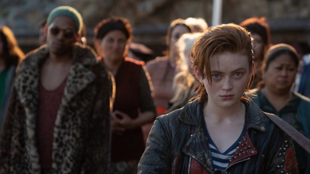 Sadie Sink with a serious expression in the foreground, wearing a leather jacket with people blurred in the background, from the show "O'Dessa."