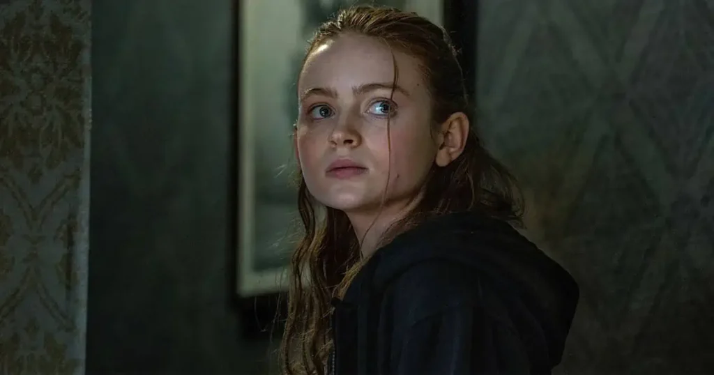 Close-up of Sadie Sink looking directly at the camera with an intense expression, wearing dark clothing.