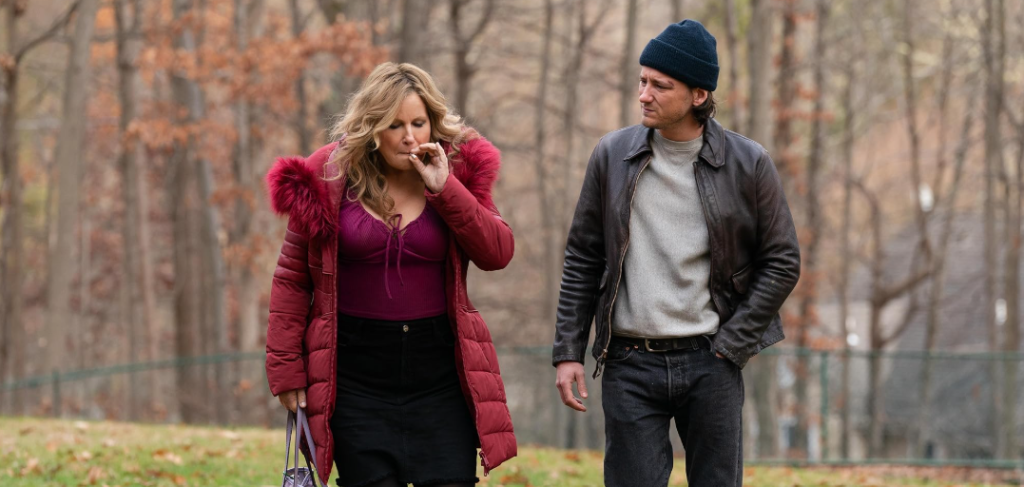 Jennifer Coolidge and Lewis Pullman are walking through a forest, with Coolidge smoking a cigarette while Pullman walks beside her.