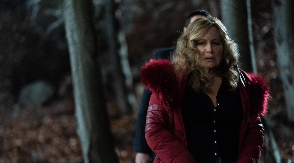 Jennifer Coolidge standing in a dark forest, dressed in a striking red outfit.