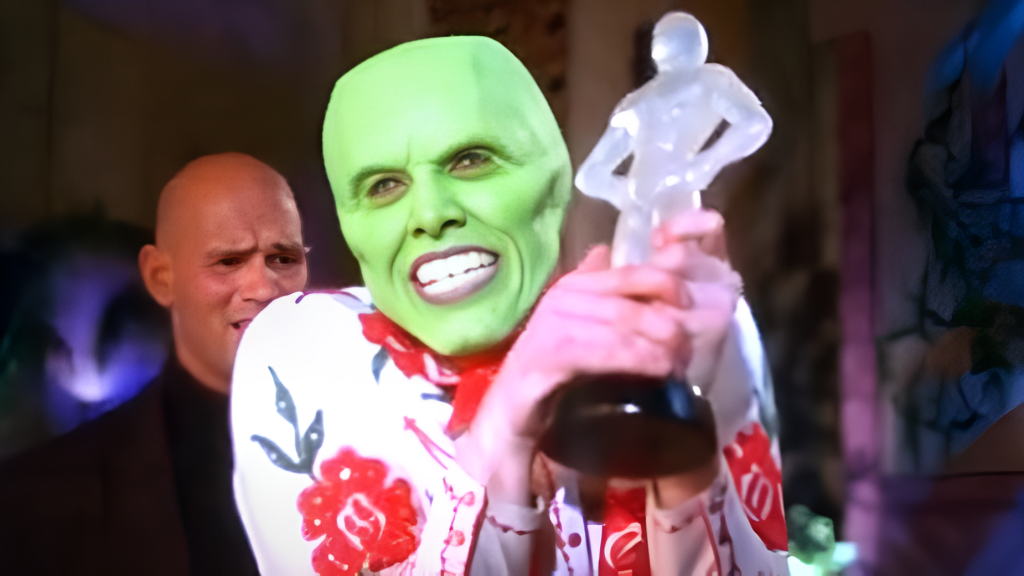 A scene from the film "The Mask" showing Jim Carrey as Mask gleefully holding up a fake Oscar statuette. Another person can be partially seen in the background.