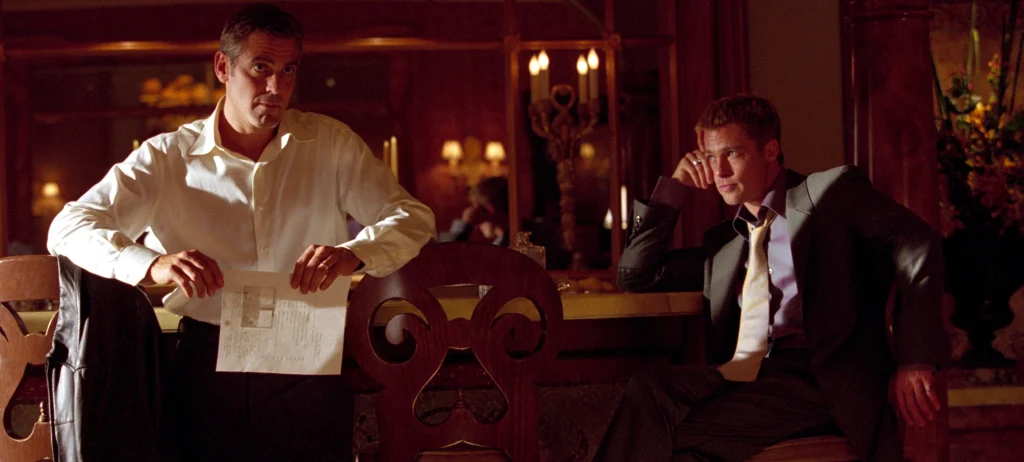 A scene from 'Ocean's 11' showing George Clooney and Brad Pitt in a richly decorated room. Clooney in a white shirt stands holding papers near an ornate wooden piece of furniture, while Pitt in formal attire sits looking thoughtful.