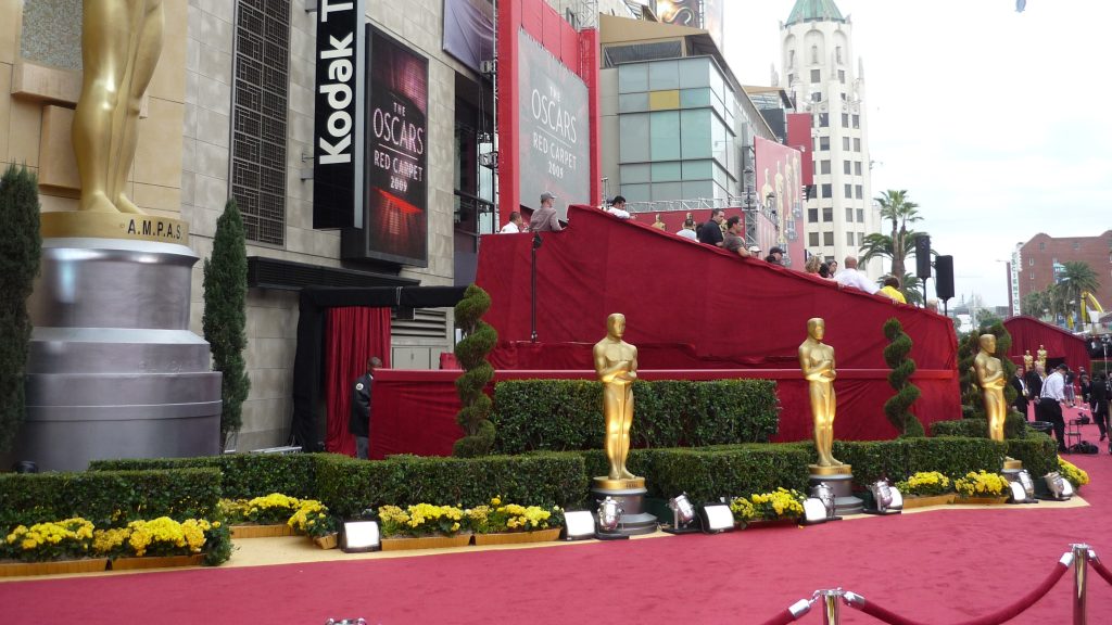 The red carpet setup for an Oscars ceremony with the iconic gold Oscar statues lining the walkway. Red carpet covers the ground with hedges, barriers, and Oscars signage visible on the Kodak Theatre building.