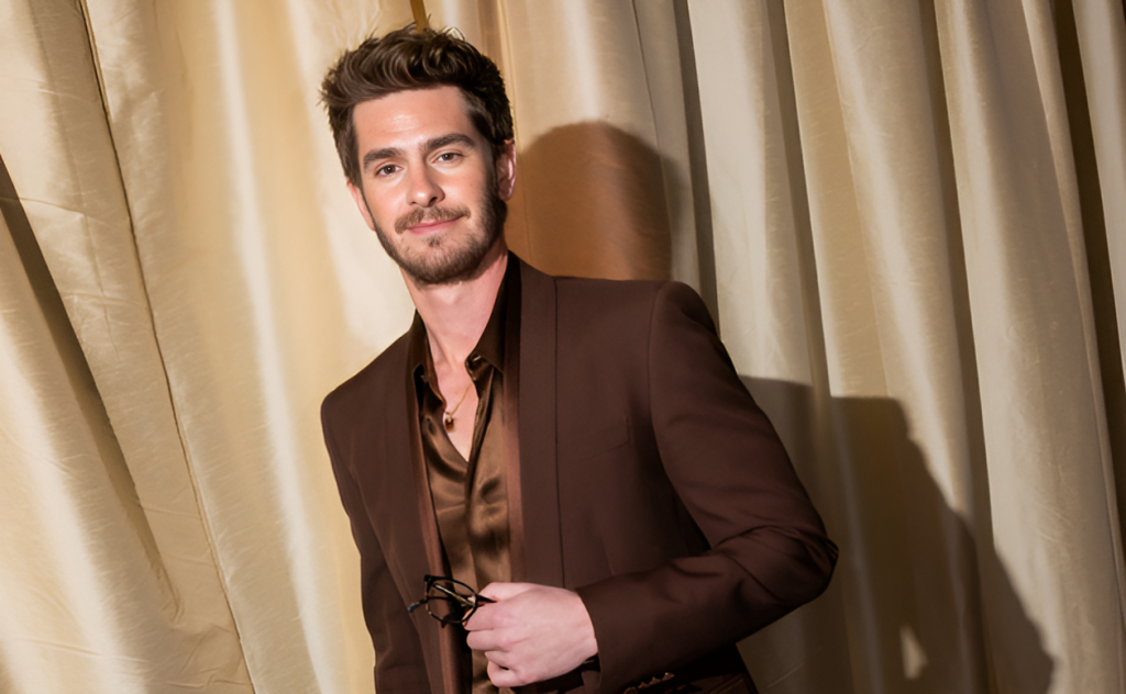 Andrew Garfield in a brown suit with a matching brown silk shirt, smiling slightly while holding glasses. He's photographed against beige curtains at the Oscar ceremony.