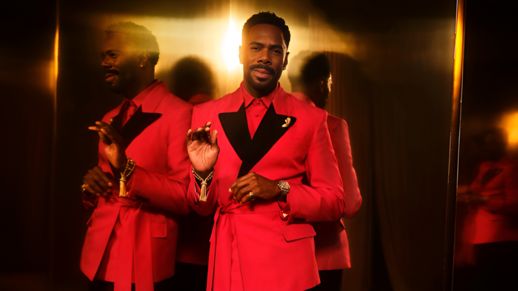 Colman Domingo in a striking red suit with black lapels, posing against a gold reflective background that creates multiple reflections.