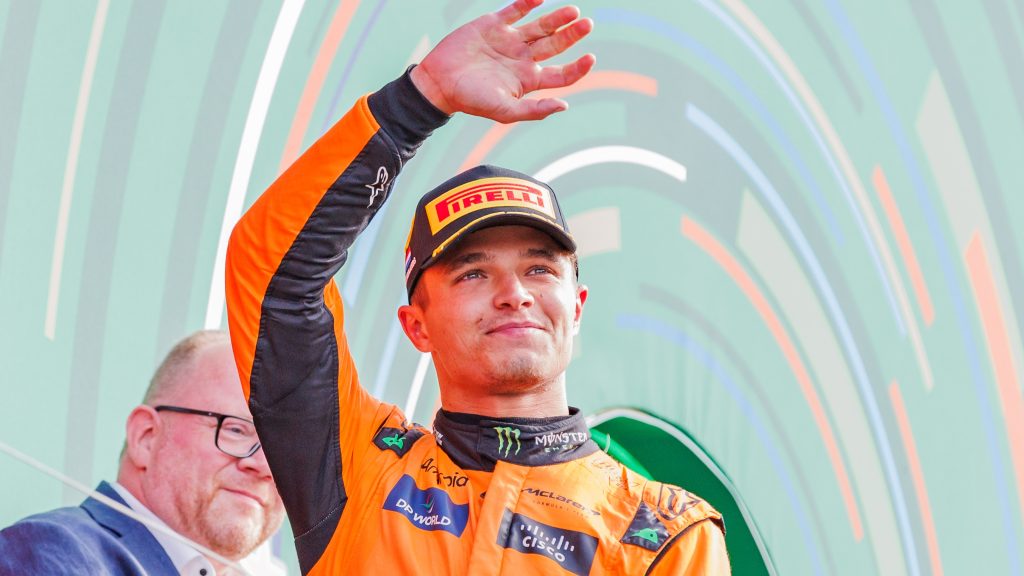 Lando Norris in orange racing suit waves to the crowd from a podium, wearing a Pirelli cap and smiling.