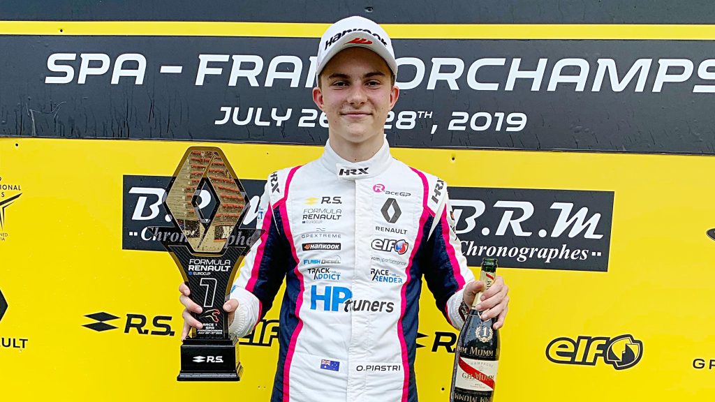 Oscar Piastri in white racing suit with pink accents holds a trophy and champagne bottle on a podium at Spa-Francorchamps in 2019.