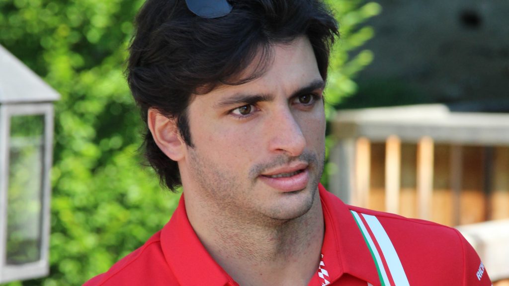 Carlos Sainz Jr. in red team polo shirt with dark hair and beard speaking or being interviewed outdoors with greenery in the background.