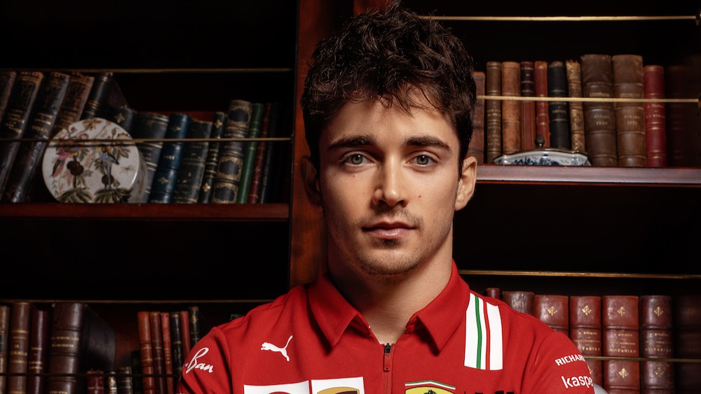 A close-up portrait of Charles Leclerc in Ferrari red team wear against a dark background with bookshelves.