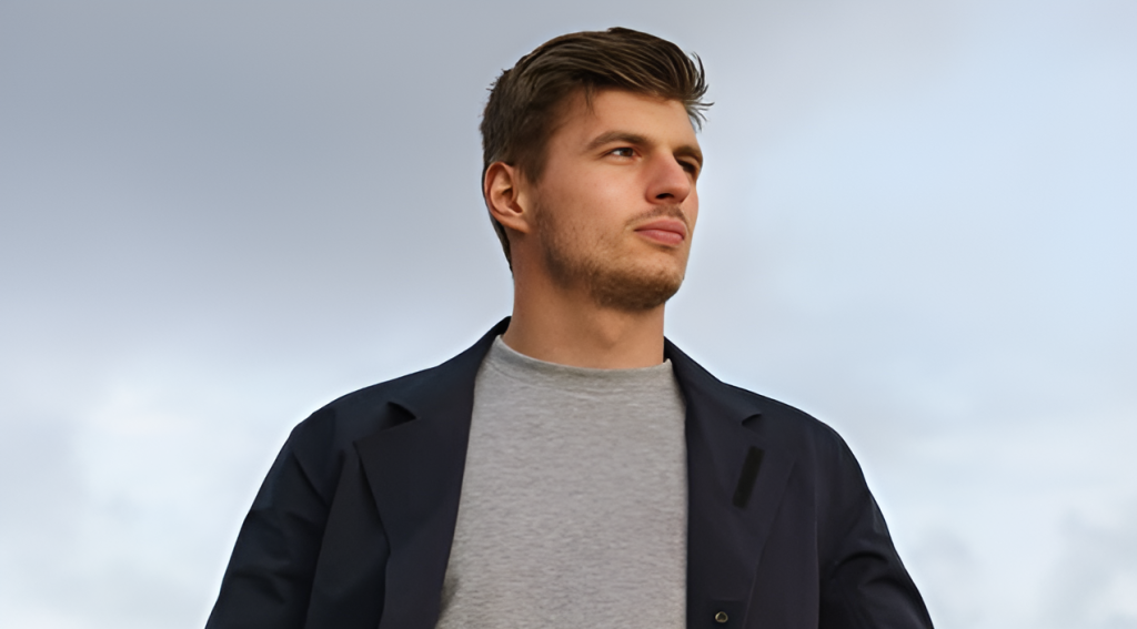 Max Verstappen in casual attire looking off into the distance against a gray background, wearing a dark blazer over a gray t-shirt.