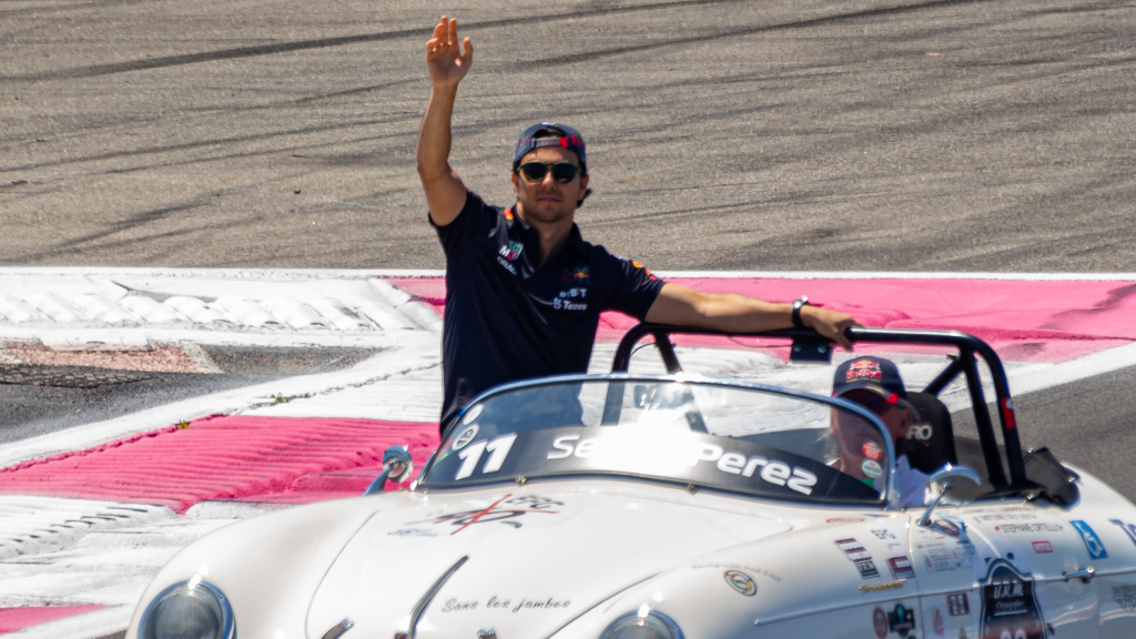 Sergio Pérez waving to fans while standing in a vintage convertible car during a parade lap or drivers' presentation.