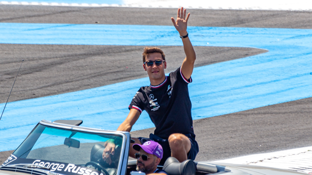 George Russell in Mercedes team gear waving to the crowd from a classic convertible car during a drivers' parade, with what appears to be a team member driving.