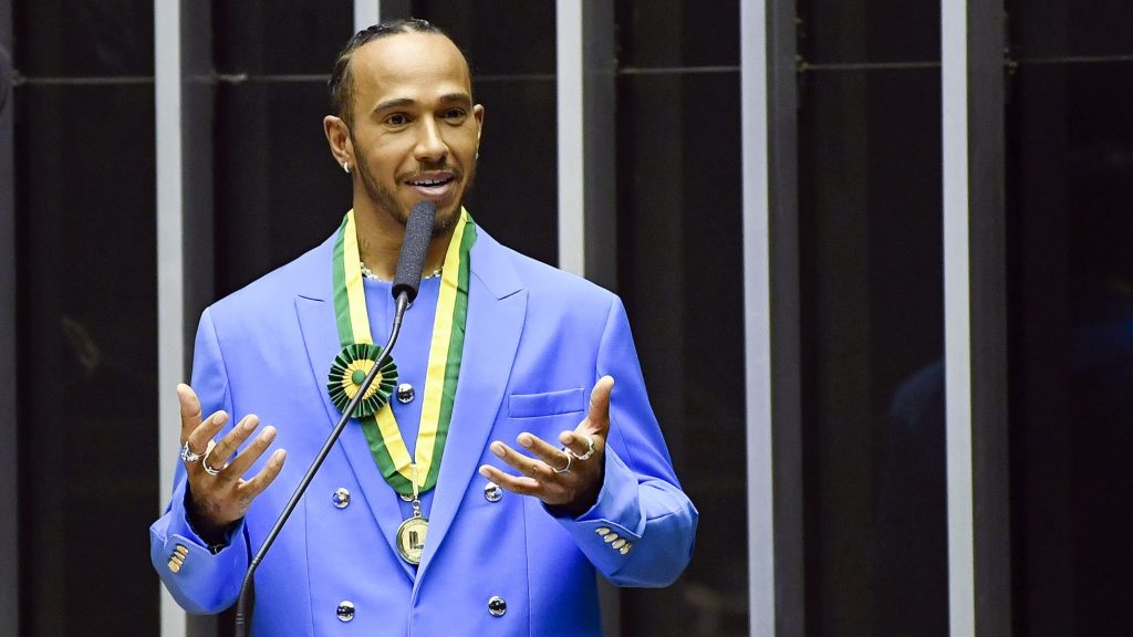 Lewis Hamilton speaking at a formal event, wearing a bright blue double-breasted suit with a medal or award featuring green and yellow colors around his neck, likely at an honorary ceremony.