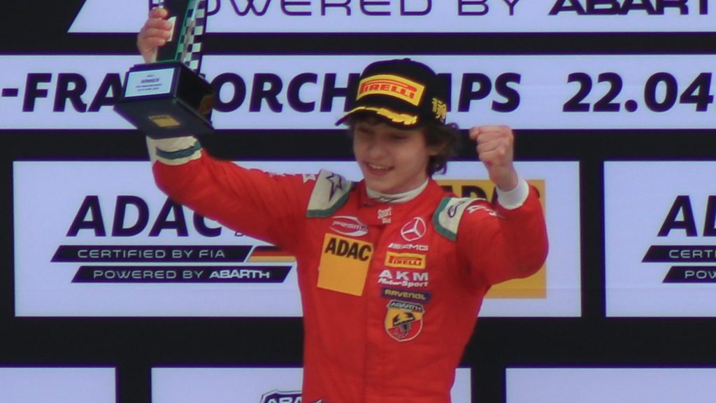 Kimi Antonelli in red racing suit celebrating on a podium, holding a trophy aloft with one fist clenched in victory.