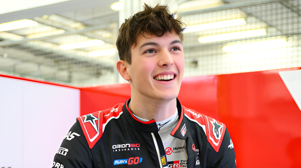 Ollie Bearman in a black and red racing suit with Haas F1 team logos smiling broadly in what appears to be a garage or team area.