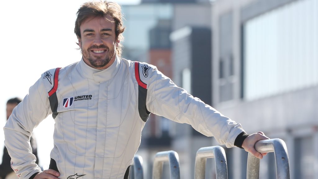 Fernando Alonso in white racing overalls with red and blue stripes, smiling at an event or testing session, with his hands resting on a barrier.