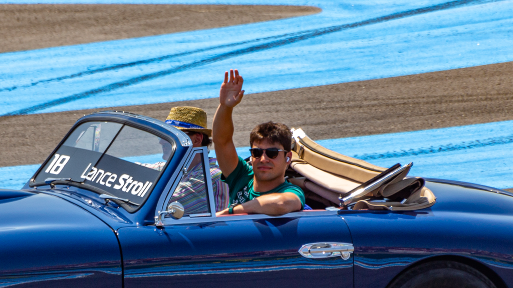 Lance Stroll waving to fans from a blue vintage convertible during a drivers' parade at a Grand Prix circuit. His name is visible on the windshield and he's wearing sunglasses and a green shirt.