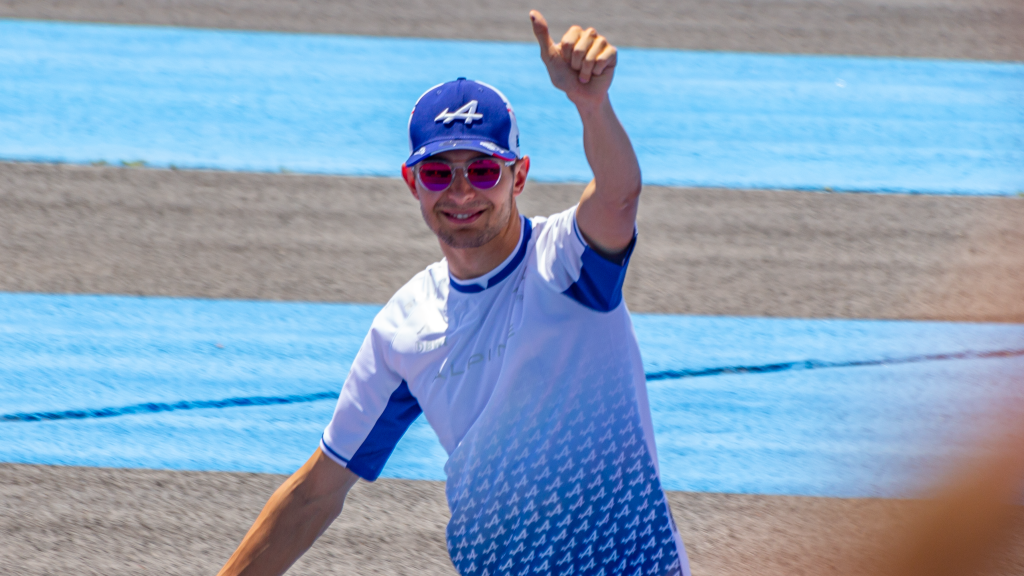 Esteban Ocon giving a thumbs up while wearing Alpine team merchandise including a blue cap with the number 44 and pink sunglasses.