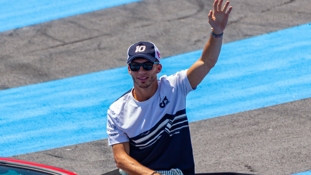 Pierre Gasly waving to spectators during a drivers' parade, wearing white AlphaTauri (now RB) team gear and sunglasses.