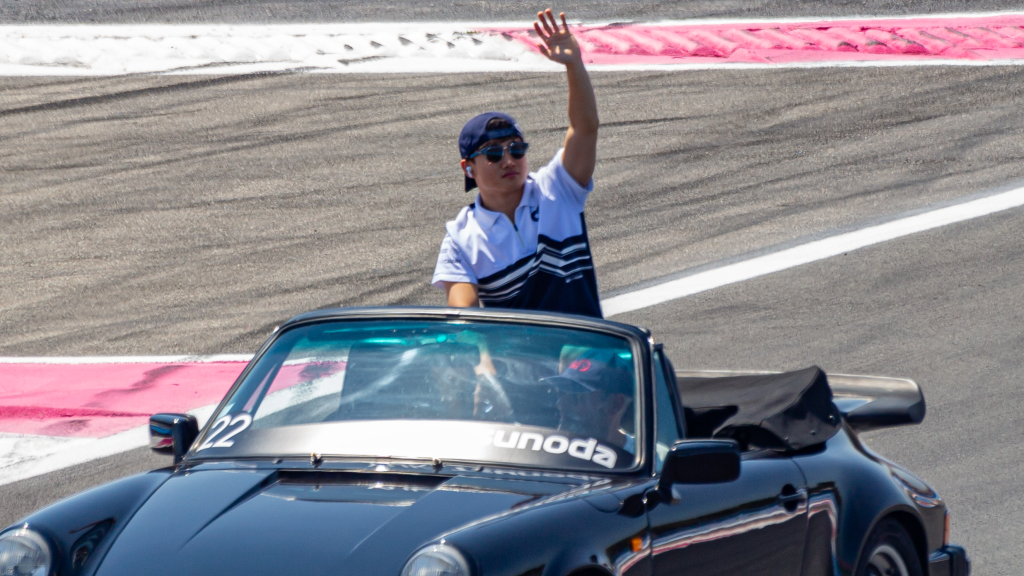 Yuki Tsunoda waving from a vintage black Porsche convertible during a drivers' parade. He's wearing white and blue team polo and a blue cap as the car travels along a circuit with red and white curbing.