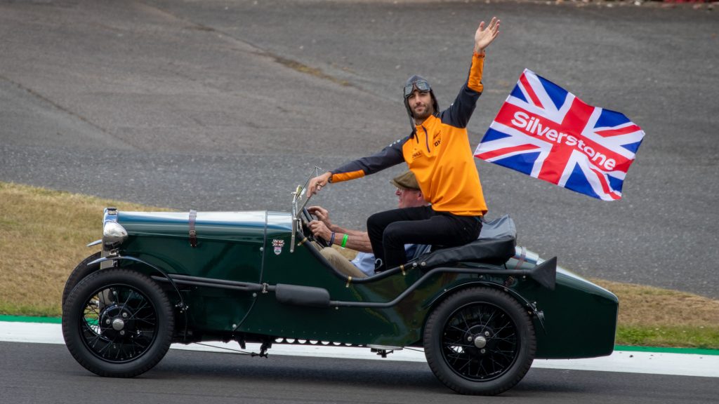 Daniel Ricciardo in McLaren orange team wear waving to fans while being driven in a vintage green sports car at Silverstone. A British flag with "Silverstone" text is visible in the background.