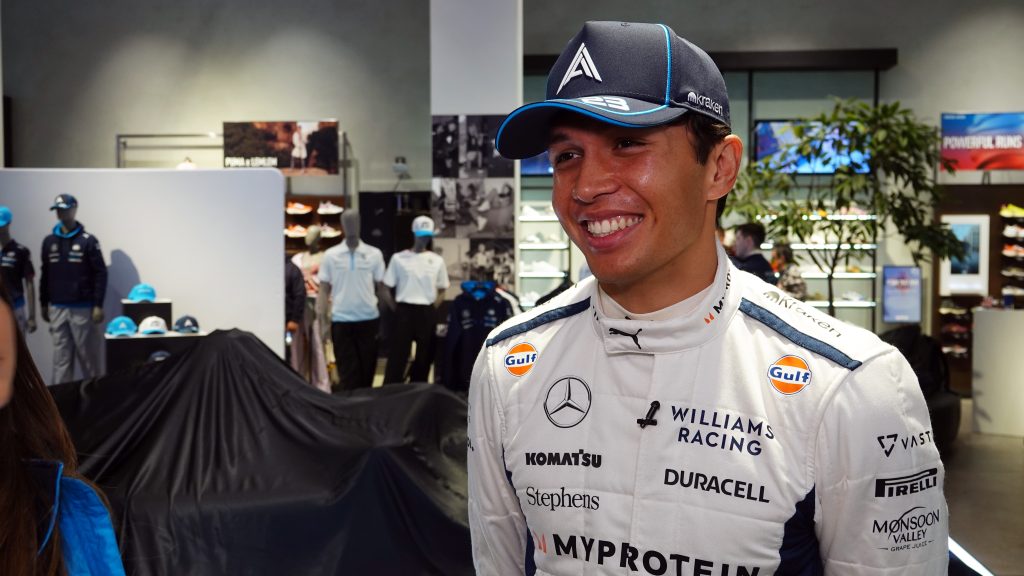 Alex Albon smiling while wearing Williams Racing team gear with a Williams cap in what appears to be a merchandise or team store with displays visible in the background.