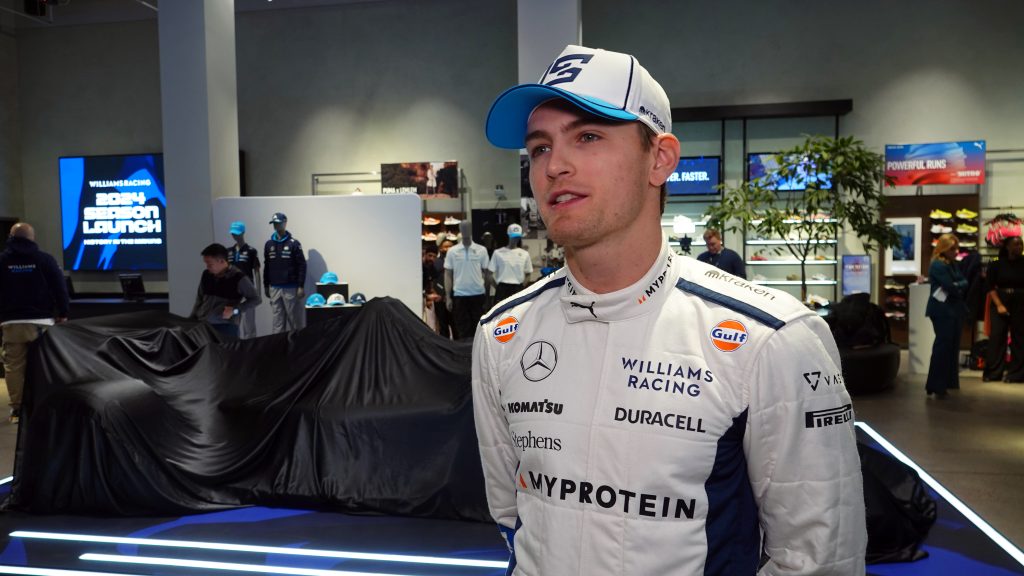 Logan Sargeant in Williams Racing team uniform and cap at what appears to be a team launch event, with a car covered under a black sheet visible behind him.