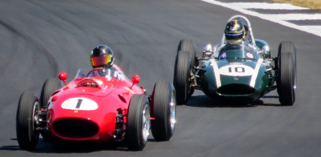 Two vintage Formula 1 cars racing on track - a red Ferrari with number 1 and a British racing green car with number 10. Both drivers are wearing distinctive period helmets typical of classic F1 eras.