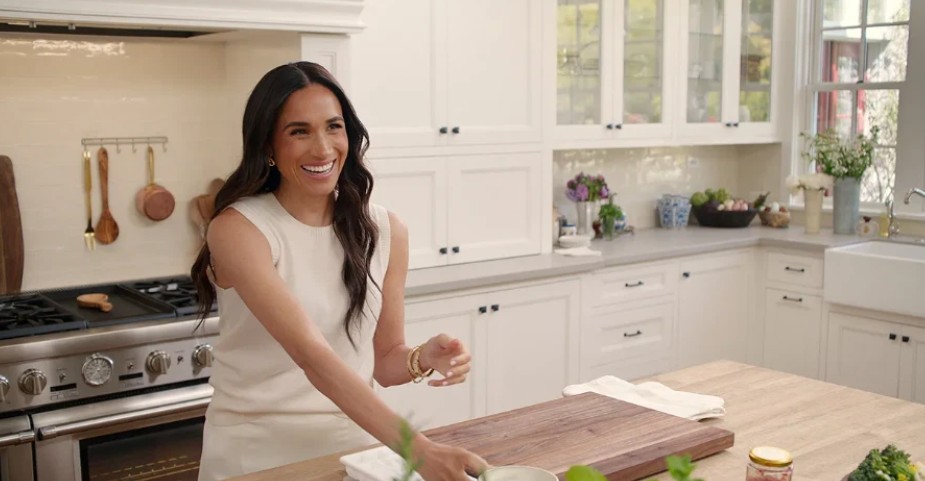 Markle in With Love, Meghan, wearing a white dress while standing in a kitchen, smiling as she prepares a meal.