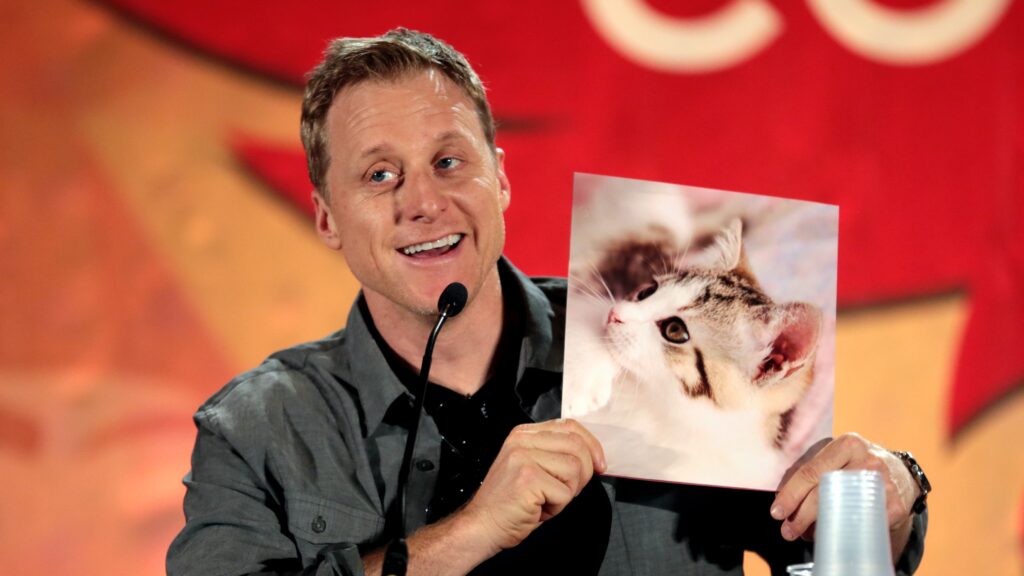 Alan Tudyk, smiling while holding up a picture of a cat. He's wearing a gray shirt and speaking into a microphone with a red backdrop behind him.