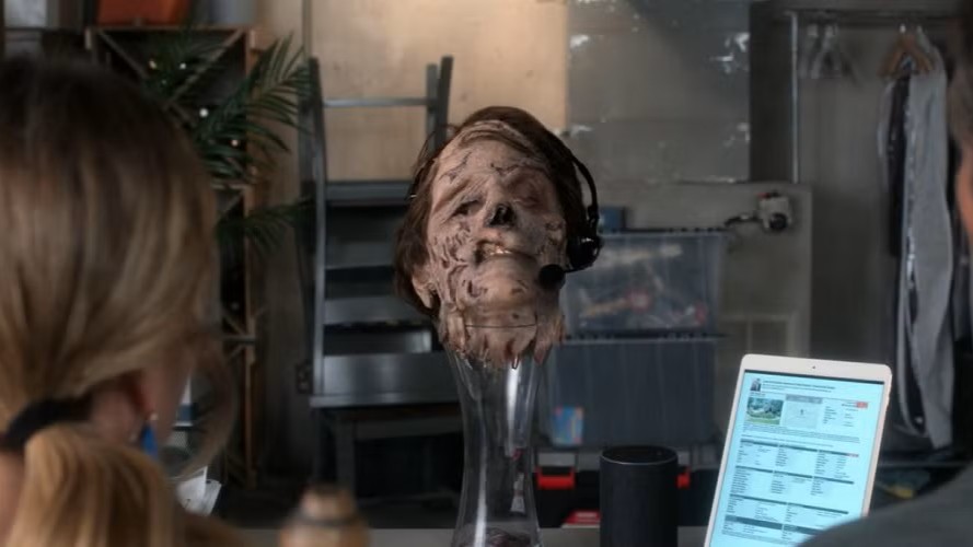 A scene from 'Santa Clarita Diet' showing Alan Tudyk as Gary West's severed, decomposing head on a stand. The character's head appears partially decayed and is wearing a headset.