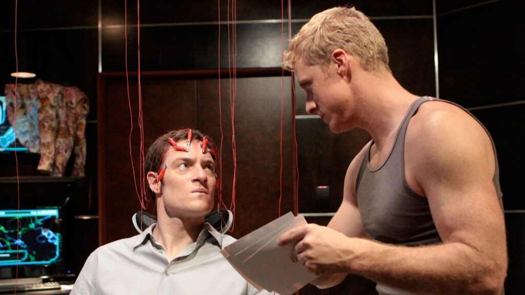 Alan Tudyk in a scene from Dollhouse where he plays Alpha. He is standing over Paul Ballard (Tahmoh Penikett) in what appears to be a testing or programming session. Paul has electrodes and wires attached to his head, and is looking up at Alpha.