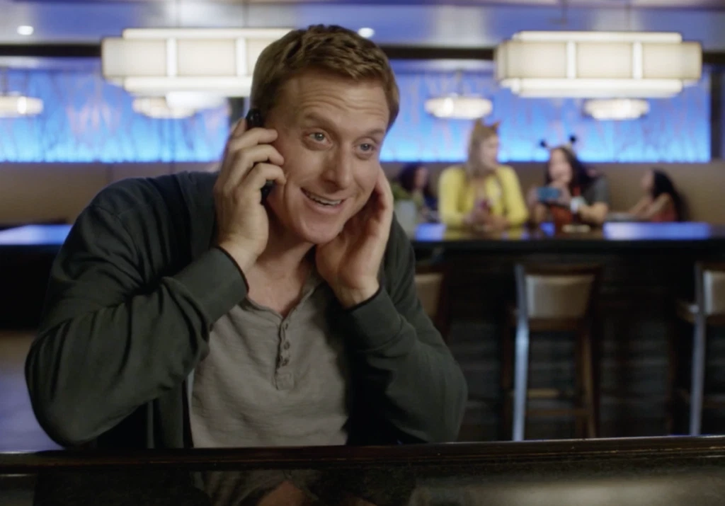 Alan Tudyk in a scene from Con Man, smiling awkwardly while on a phone call in what appears to be a restaurant or diner.