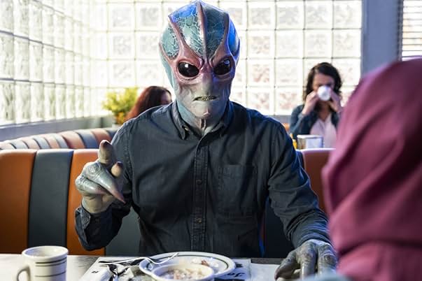 Alan Tudyk in makeup and costume as Harry Vanderspeigle from Resident Alien, showing his alien appearance with blue-tinted skin and large, dark eyes. He's seated at a diner booth.