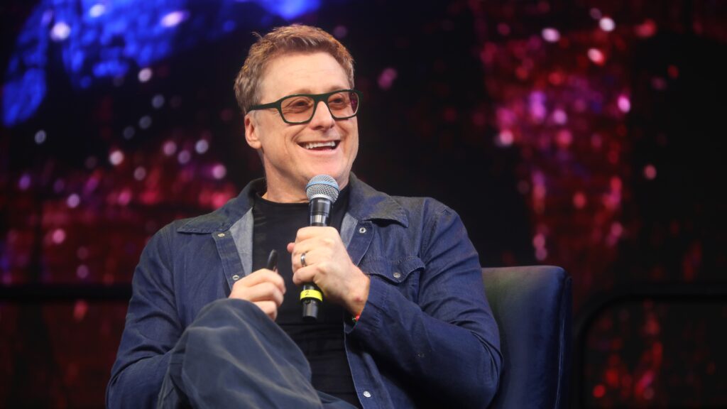 Alan Tudyk smiling broadly while speaking into a microphone. He's wearing glasses and a blue denim shirt, seated against a colorful background with blue and red lighting.