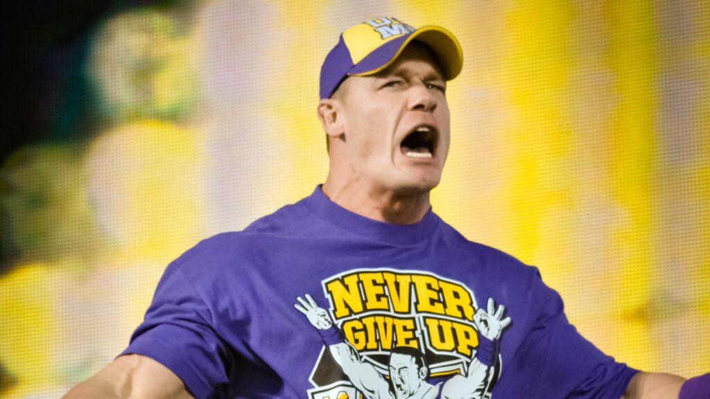 John Cena in his purple and yellow "Never Give Up" merchandise, shouting energetically during his entrance. He's wearing a WWE cap.