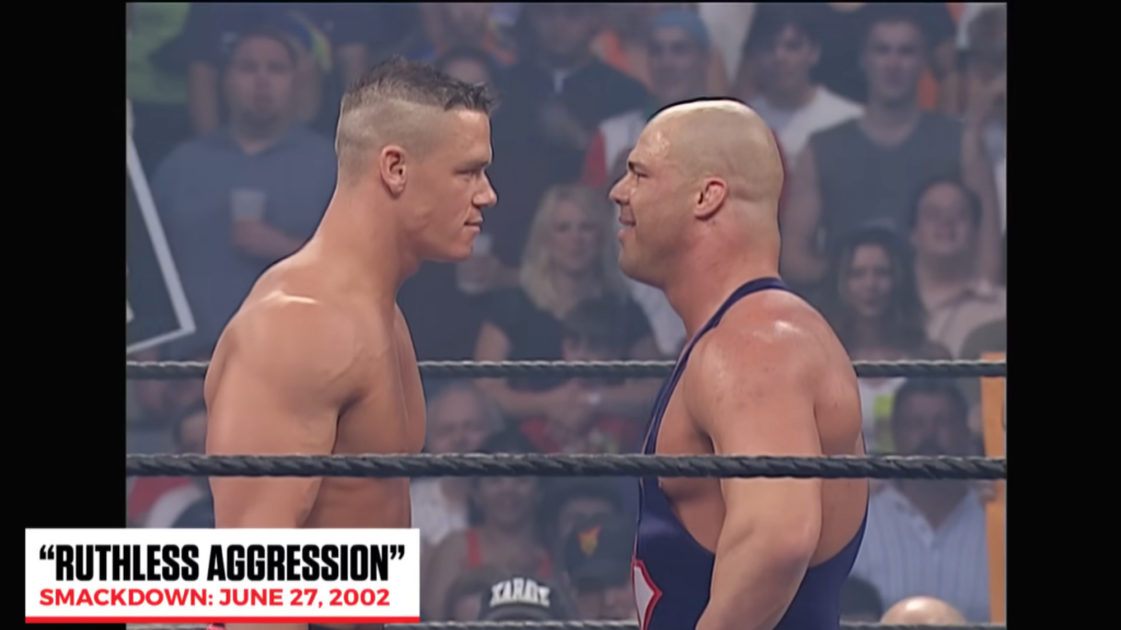 John Cena facing Kurt Angle in the ring during his WWE debut on SmackDown, June 27, 2002. The image shows the famous "Ruthless Aggression" moment when rookie Cena first challenged the Olympic gold medalist.