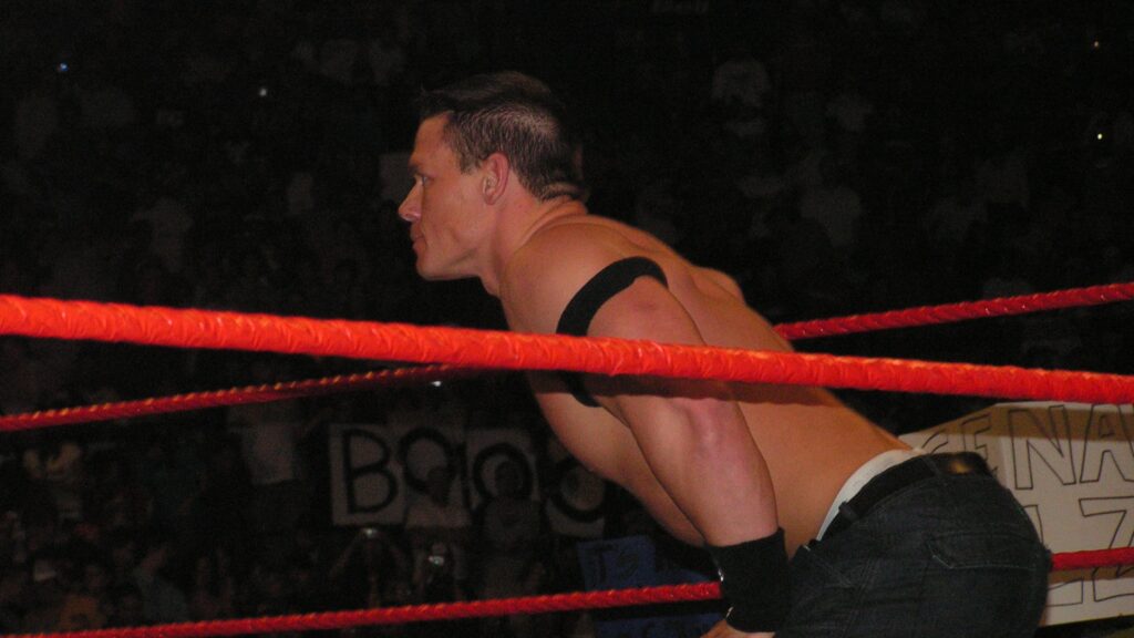 A young John Cena leaning on the ring ropes during an early WWE match. He has short hair and is wearing minimal wrestling gear.