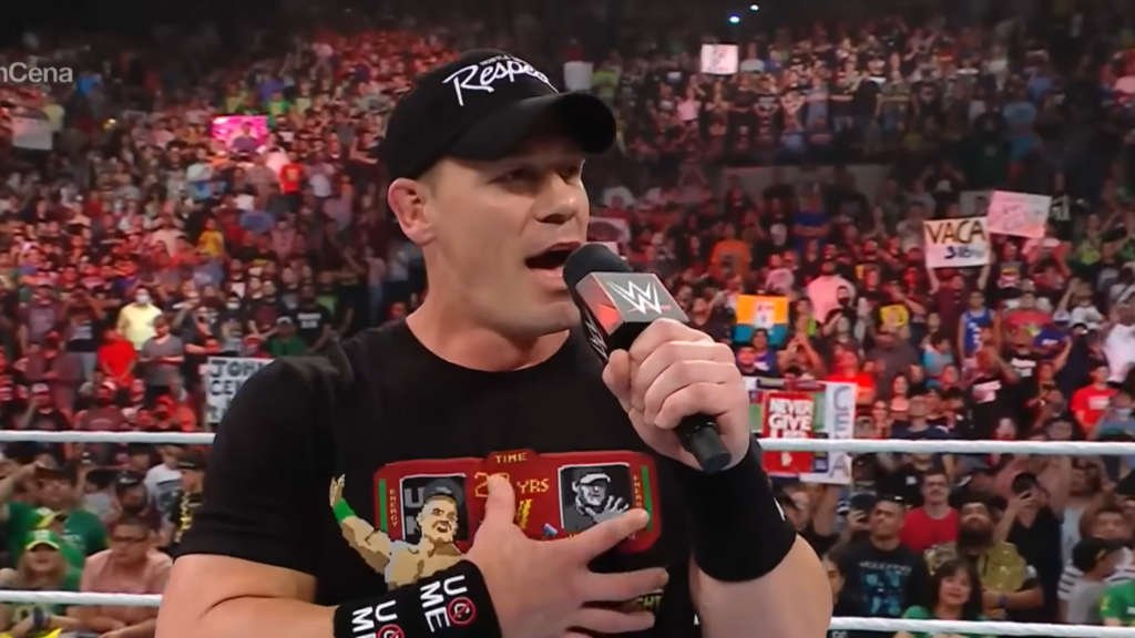 John Cena in black attire with his "Respect" cap, holding a microphone in a WWE ring. He's wearing his signature wristbands and a t-shirt featuring his logo. A passionate crowd can be seen in the background.