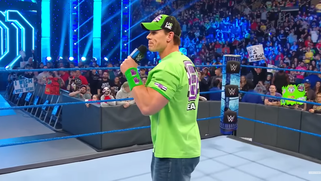 John Cena in a bright green t-shirt and cap, standing in a WWE ring with a microphone. The crowd can be seen in the background.