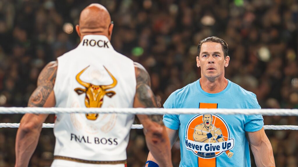 The Rock (with "ROCK" visible on the back of his vest) facing off against John Cena in a blue "Never Give Up" t-shirt in a WWE ring.