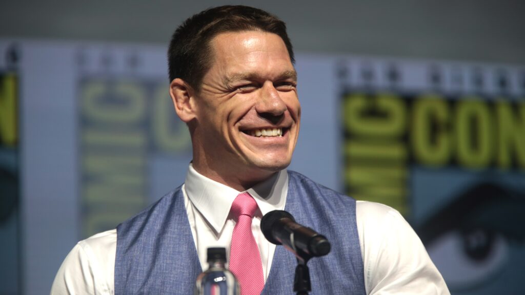 John Cena in a suit and pink tie smiling while speaking at a microphone during a Comic-Con panel.